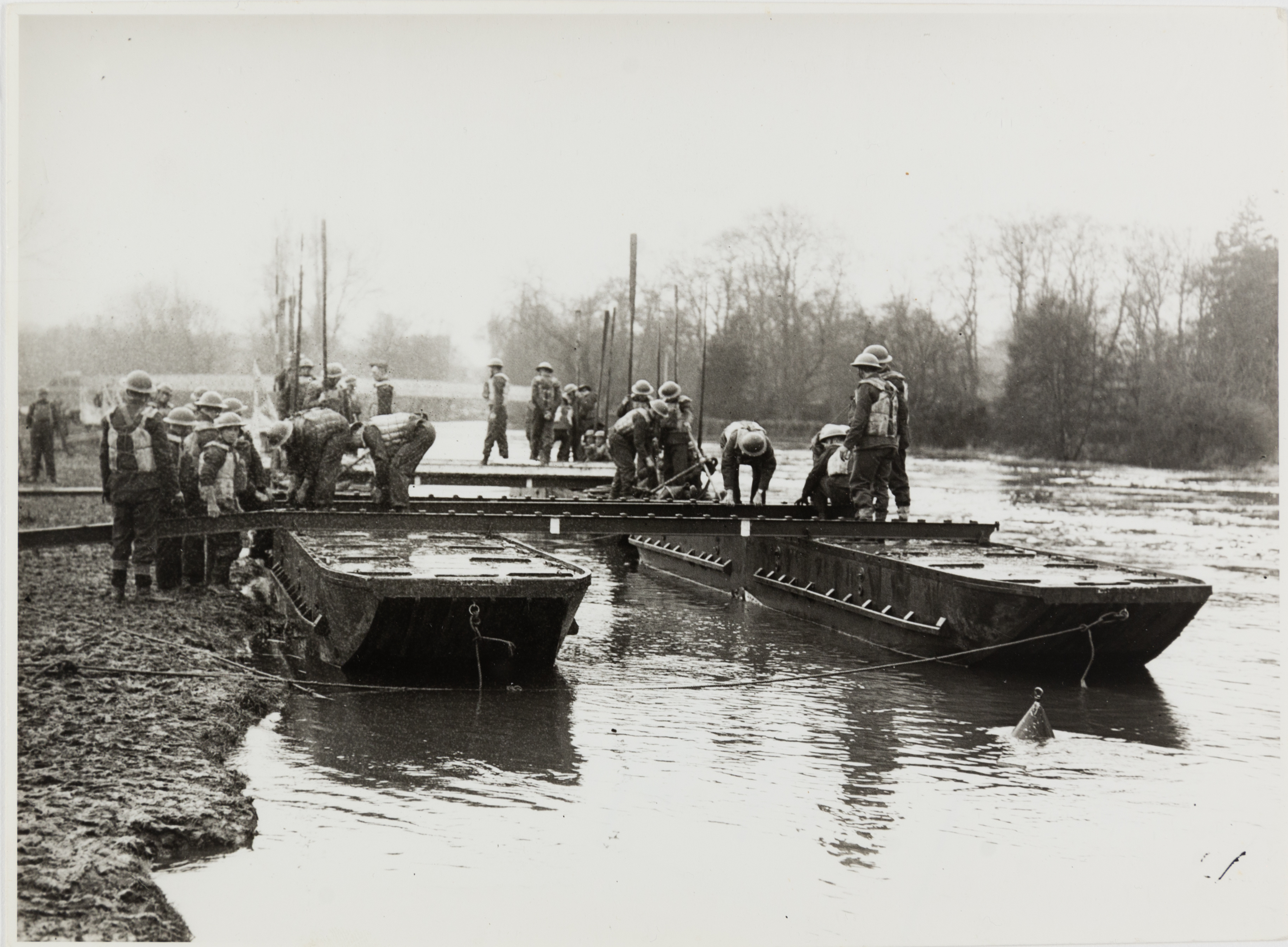 Black and white photograph of several soldiers standing and sitting on moored landing crafts in the Thames. There is a metal frame between two crafts with soldiers are attending to.